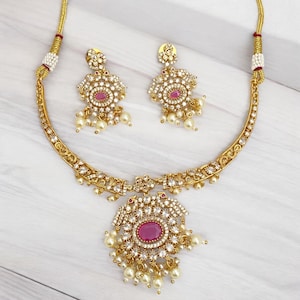 May include: A gold necklace and earrings set with a pink stone and white accents. The necklace has a pendant with a round pink stone surrounded by white stones and a fringe of white pearls. The earrings have a similar design with a smaller pink stone.