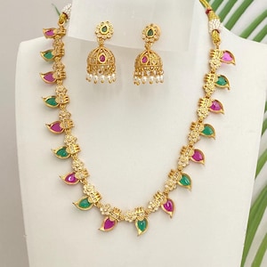 May include: Gold-toned necklace and earring set. The necklace features alternating green and pink leaf-shaped pendants with small white accents. The earrings are bell-shaped with a floral design and small white pearl dangles.