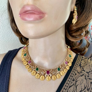 May include: A gold necklace with a coin design and red and green gemstones. The necklace is on a mannequin wearing a black top and a gold and black patterned sari.