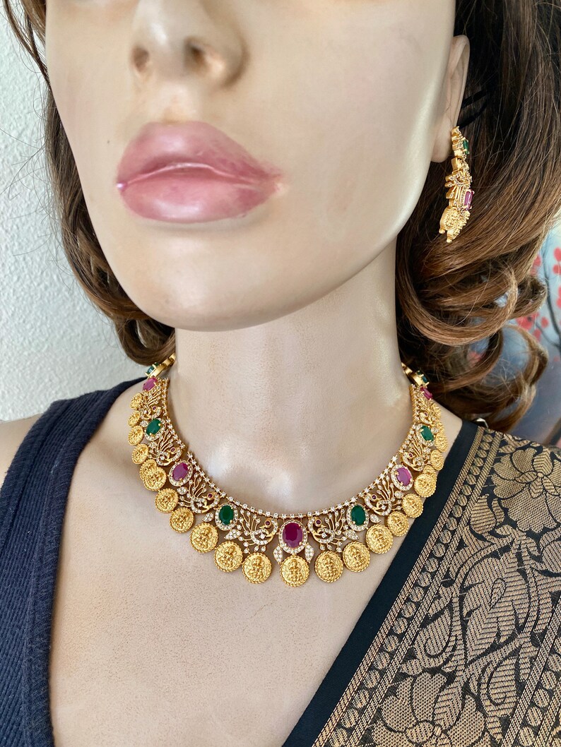 May include: A gold necklace with a coin design and red and green gemstones. The necklace is on a mannequin wearing a black top and a gold and black patterned sari.