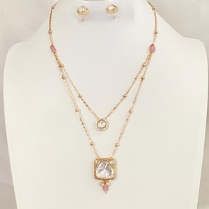 May include: A gold-toned layered necklace featuring two chains with small metallic beads and pink oval accents. The upper chain has a small circular pendant, while the lower chain holds a larger square pendant with a central clear stone and pink beads.