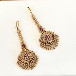 May include: A pair of gold-toned metal earrings featuring an elongated design with a hook closure. The base of each earring displays a circular motif with embedded red and green stones, accented by small dangling gold beads and peacock motifs.
