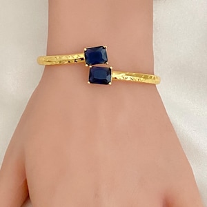 May include: Gold-tone cuff bracelet featuring two square, dark blue gemstones. The bracelet has a textured, hammered finish. The gemstones are set in a square gold setting. The bracelet is worn on a wrist.