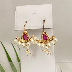 May include: Gold-toned earrings with a teardrop-shaped, magenta-colored stone at the center. The earrings feature a curved design with small, white pearls and a decorative gold border. The earrings are displayed on a white background.