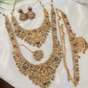 May include: A gold-toned jewelry set featuring two necklaces, a pair of jhumka earrings, a maang tikka, and a waist belt. Each piece is intricately detailed with embossed deity figures, small red and green enamel accents, and dangling beads.