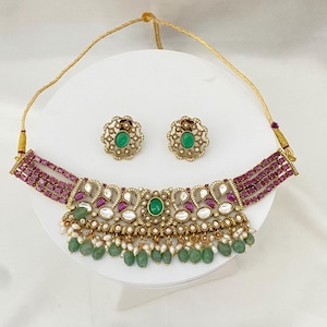 May include: A gold-toned choker necklace and matching stud earrings set. The necklace features rows of oval-cut magenta stones, a central green stone, white accents, and dangling green beads with small white pearls. The earrings have a matching design.