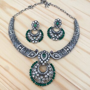 May include: A silver necklace with a peacock design and green and white stones. The necklace has a matching pair of earrings with a similar design.