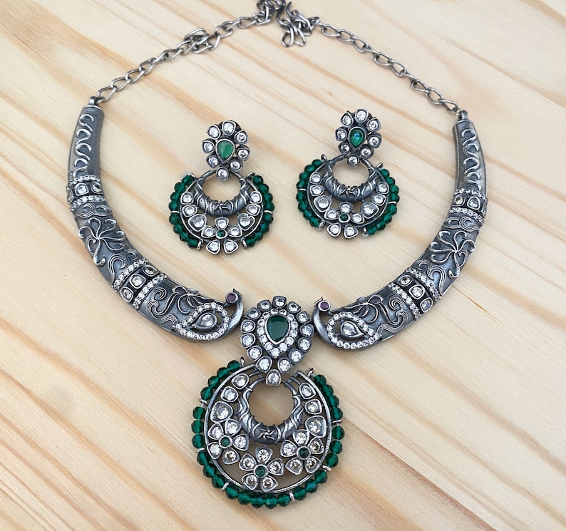 May include: A silver necklace with a peacock design and green and white stones. The necklace has a matching pair of earrings with a similar design.