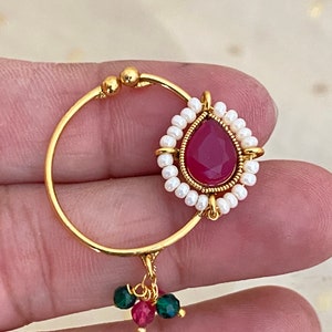 May include: A gold-colored nose ring featuring a teardrop-shaped, deep red gemstone surrounded by small white pearls. The ring has a circular design with a small cluster of green and red beads hanging from the bottom.