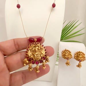May include: A gold-toned pendant necklace and matching earrings set. The pendant features a detailed deity design with red gemstone accents. The earrings have a similar design. The necklace has red bead accents.