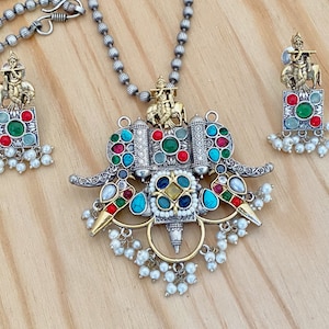 May include: A silver and gold necklace with a multi-colored pendant featuring an elephant design and a small figure of a person playing a flute. The pendant is adorned with pearls and colorful stones. Matching earrings with a similar design are also included.