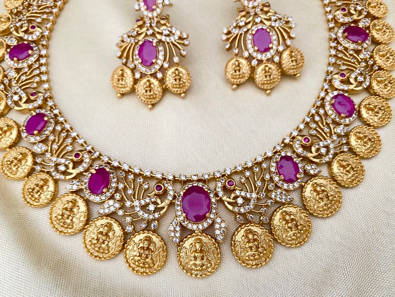 May include: A gold necklace with a coin design and pink gemstones. The necklace is paired with matching earrings.