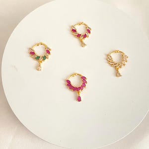 May include: Four gold-toned nose rings with gemstone accents. Each ring features a circular design with a cluster of gemstones in various colors, including pink, green, and clear, and a dangling teardrop-shaped gem.