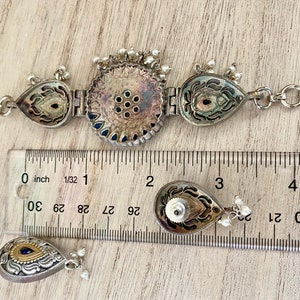 May include: A silver and blue necklace with a central round pendant and two teardrop pendants. The necklace is measured against a ruler with markings in inches.