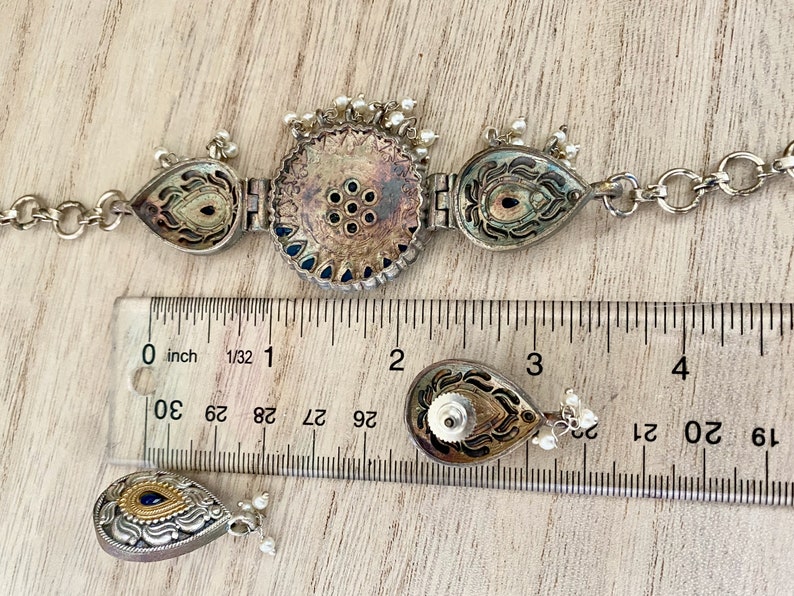May include: A silver and blue necklace with a central round pendant and two teardrop pendants. The necklace is measured against a ruler with markings in inches.