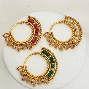 May include: Three gold-toned hoop earrings with intricate detailing. Each earring features a crescent-shaped design adorned with gemstones. One has red beads, another has pearls, and the third has green beads. The earrings have a teardrop-shaped design.