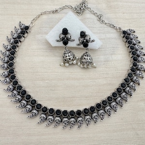 May include: A silver-toned necklace and matching earrings set. The necklace and earrings feature black gemstones and ornate, leaf-shaped metalwork. The earrings have a bell-shaped design with small white beads. The jewelry set is displayed on a light-colored surface.