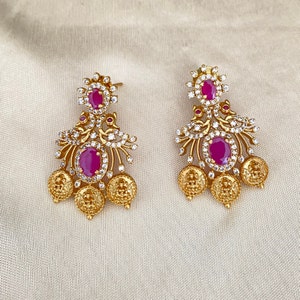 May include: A pair of gold earrings with pink stones and white accents. The earrings have a floral design and dangle from a small loop.