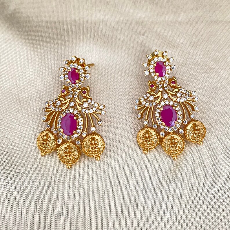 May include: A pair of gold earrings with pink stones and white accents. The earrings have a floral design and dangle from a small loop.