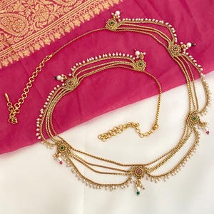 May include: A gold-toned waist chain featuring multiple delicate box chains draped in layers. The design includes circular floral motifs accented with small green and red stones, finished with dangling white pearl beads along the lower edges.