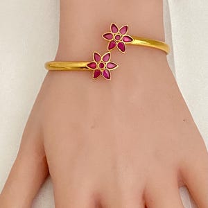 May include: A gold-colored bangle bracelet featuring two flower-shaped designs with pink gemstone centers. The bracelet is worn on a wrist, showcasing its elegant and delicate floral details. The jewelry is set against a soft, neutral background.