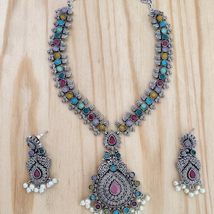 May include: A silver necklace with a pendant featuring a multi-colored floral design and a pink stone. The necklace is paired with matching earrings with pearl accents.