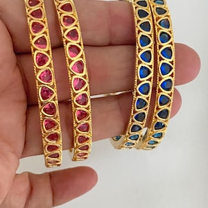 May include: Gold-toned bangles with colorful gemstones. Two bangles feature pink gemstones, while the other two showcase blue gemstones. The gemstones are set in a repeating pattern along the bangles.