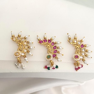 May include: Three ornate gold-toned earrings with a peacock design. Each earring features clear and ruby-colored gemstones, small pearls, and a central peacock motif. The earrings are displayed on a white surface.