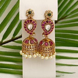 May include: Gold-toned earrings with a floral top, teardrop-shaped center, and bell-shaped bottom. The earrings feature red and clear gemstones, with a pearl fringe. The earrings are displayed on a white stand against a backdrop of green leaves.