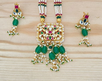 May include: A gold necklace with a square pendant featuring intricate floral designs and green and pink gemstones. The pendant is attached to a string of white pearls and green beads. The necklace comes with matching earrings with green and pink gemstones.
