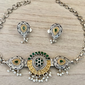 May include: A silver and gold necklace with a green stone pendant and matching earrings. The necklace has a chain link design and the pendant is surrounded by intricate details.