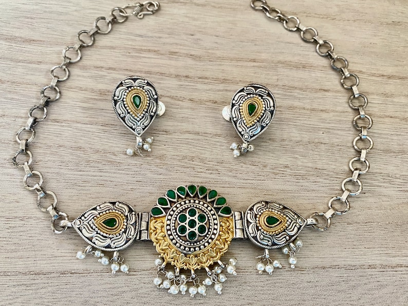 May include: A silver and gold necklace with a green stone pendant and matching earrings. The necklace has a chain link design and the pendant is surrounded by intricate details.