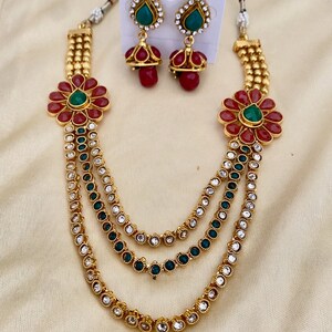 Puede incluir: Un collar de oro de varias capas con detalles de flores rojas y verdes y pendientes a juego. El collar tiene tres capas de cadena de oro con piedras transparentes y verdes.
