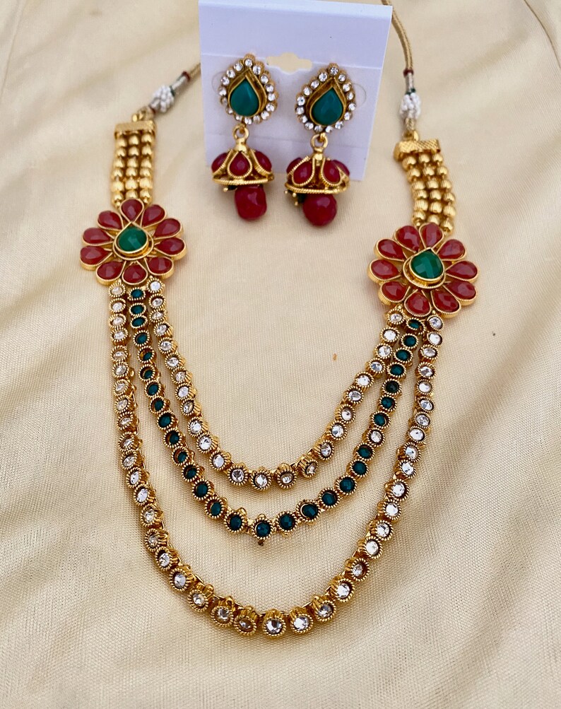 Puede incluir: Un collar de oro de varias capas con detalles de flores rojas y verdes y pendientes a juego. El collar tiene tres capas de cadena de oro con piedras transparentes y verdes.
