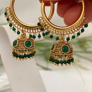 May include: Gold-toned hoop earrings with intricate detailing. Each earring features a series of small green beads and a bell-shaped charm adorned with green gemstones. The earrings are held up by a hand.