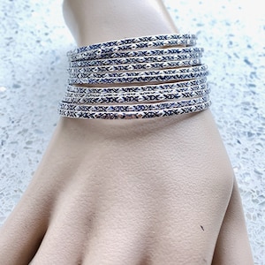 May include: A stack of silver bangles with a repeating blue and white geometric pattern. The bangles are stacked on a light-toned surface, showcasing the intricate design and metallic sheen.