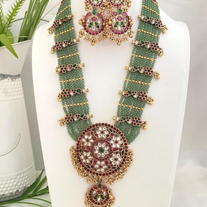 May include: A multi-strand necklace featuring small sage green beads and gold-toned spacers with red and green floral accents. A large circular pendant with red, green, and white stones hangs at the center, accompanied by matching round drop earrings.
