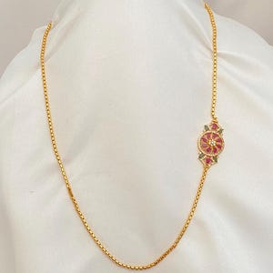 May include: A gold-colored necklace with a floral pendant. The pendant features a central cluster of red gemstones surrounded by smaller green and red stones. The necklace is displayed on a white fabric background.