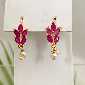 May include: A pair of gold-colored earrings. Each earring features a cluster of fuchsia-colored, marquise-cut gemstones arranged in a leaf-like pattern. Small, white pearls dangle from the bottom of each earring, adding a delicate touch.