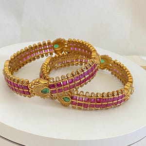 May include: Two gold-toned bangles featuring rows of square-cut magenta stones and small clear accent stones. Each bangle includes a decorative metal element shaped like a peacock head with a single oval green stone set in the center.