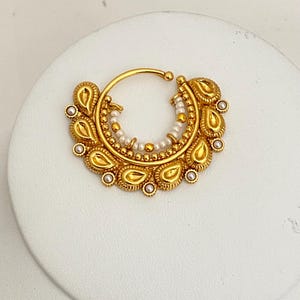 May include: A gold-colored nose ring with a circular design. The ring features a crescent-shaped pendant adorned with teardrop-shaped elements and small pearls. The design includes a central row of pearls and intricate detailing.