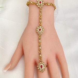 May include: Gold-toned hand chain jewelry featuring a ring, a bracelet, and a chain connecting them. The jewelry has a floral design with red and white gemstones. The bracelet and ring are connected by a delicate chain.