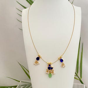 May include: A delicate gold-toned necklace featuring blue teardrop-shaped pendants and small white pearl accents. The necklace also includes a central pendant with a peacock design and a green bead. The jewelry is displayed on a white mannequin.