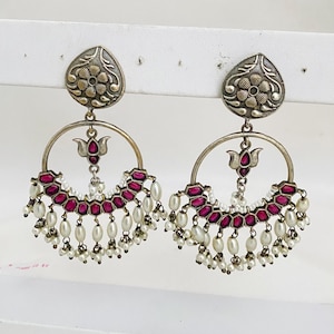 May include: A pair of silver-toned earrings with a floral design at the top and a half-circle design. The half-circle is adorned with red stones and white pearls. The earrings have a drop design.