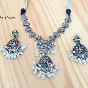 May include: A silver necklace and earrings set with a floral design. The necklace features a pendant with green, pink, and white stones. The earrings have a similar design with green and pink stones. The set is adorned with white pearls.