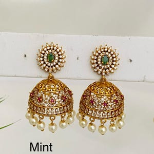 May include: Gold-toned earrings with a floral design. Each earring features a green gemstone center, surrounded by clear crystals. The lower portion has a bell shape with red and clear accents, and dangling pearls. The word "Mint" is visible.