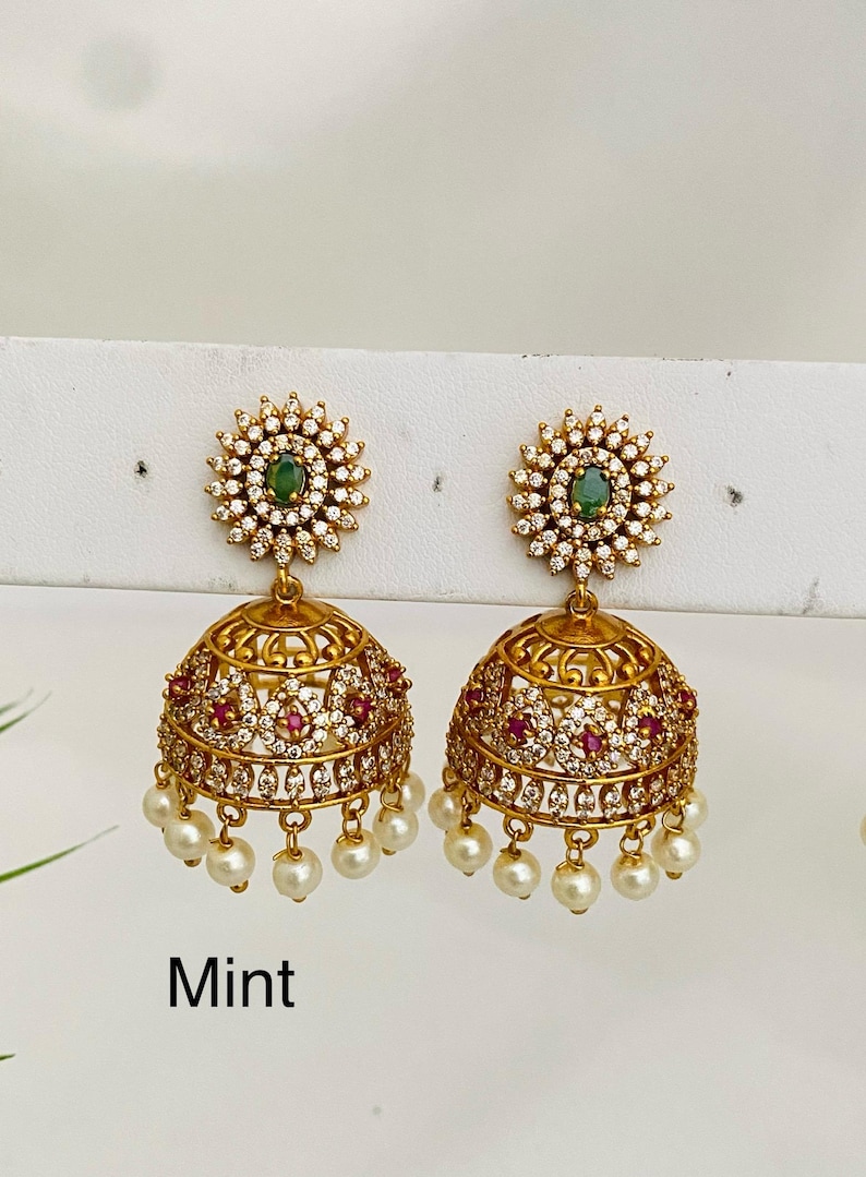May include: Gold-toned earrings with a floral design. Each earring features a green gemstone center, surrounded by clear crystals. The lower portion has a bell shape with red and clear accents, and dangling pearls. The word "Mint" is visible.