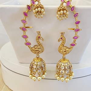 May include: A pair of gold-tone ear cuffs featuring a peacock motif with pink marquise-cut stones along the arch. Each piece includes a dangling bell-shaped jhumka charm encrusted with clear stones and finished with small white pearl-like beads.