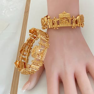 May include: A pair of gold-toned hinged bracelets featuring intricate relief carvings of figures, elephants, and a central palanquin structure. The metalwork displays detailed textures and a polished finish, with a clasp mechanism visible on the side.