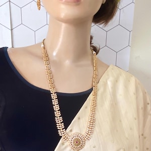 May include: Gold-toned necklace and matching earrings. The necklace features a long chain with leaf-like designs and a central pendant with a red stone. The earrings are small hoops. The jewelry is displayed on a mannequin.